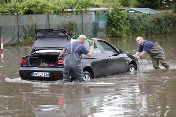 Zwei Personen ziehen ein Auto aus dem Überschwemmungsbereich. Das Wasser steht kniehoch auf der Straße.