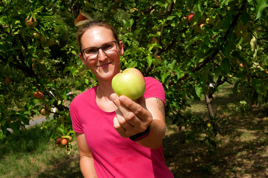 Eine Frau zeigt einen Apfel, im Hintergeund Apfelbäume.