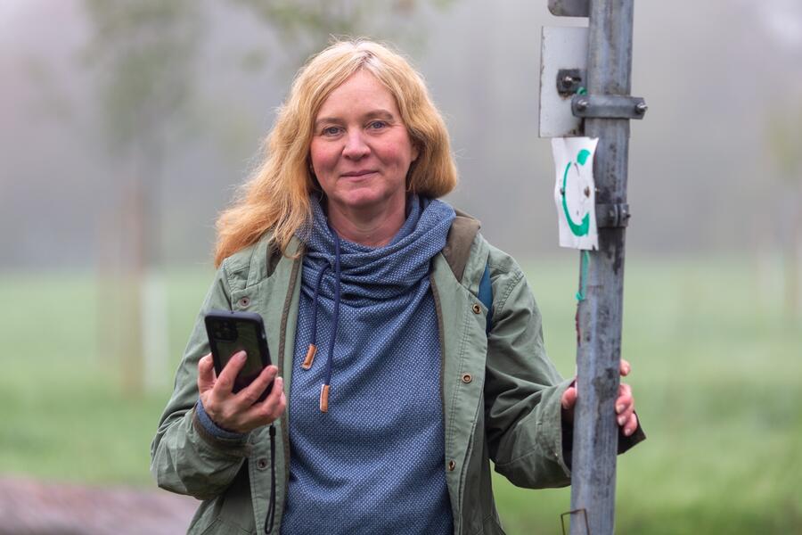 Eine Frau steht an einem Schild mit dem Symbol der Hessischen Apfelwein- und Obstwiesenroute. In der Hand hält sie ien Smartphone.