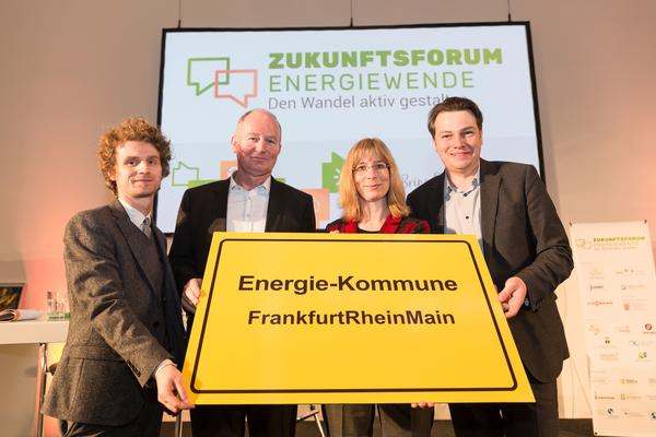 Das Bild zeigt Herrn Voll, Frau Dr. Schröder-Goga vom Regionalverband und Herrn Vohrer von der Agentur für erneuerbarer Energie wie sie den Preis, ein gelbes Schild mit dem Aufdruck "Energie-Kommune FrankfurtRheinMain", der Kamera entgegenhalten.