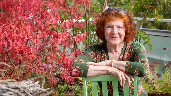 Eine Frau mit rotem Haar sitzt auf einem Balkon. Um sie herum stehen Pflanzen im Herbstlaub.