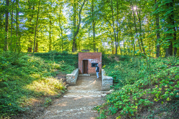 In einem lichten Wald ist ein gemauerter Kellereingang vor einem H&uuml;gel. Ein Weg f&uuml;hrt zum Eingang. Eine Person steht vor dem Geb&auml;ude.