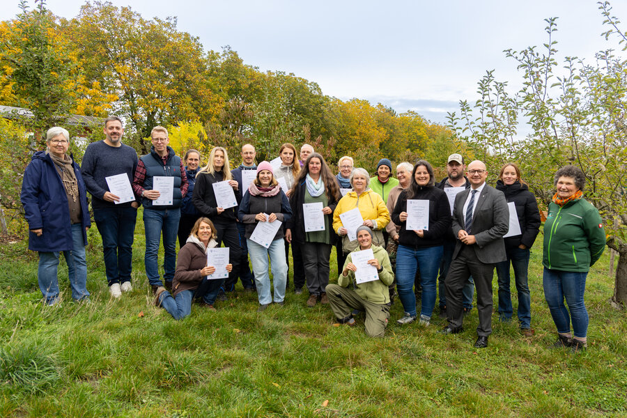Eine Gruppe von &uuml;ber 20 Menschen steht in einer herbstlichen Umgebung und halten ihre Urkunden in den H&auml;nden.