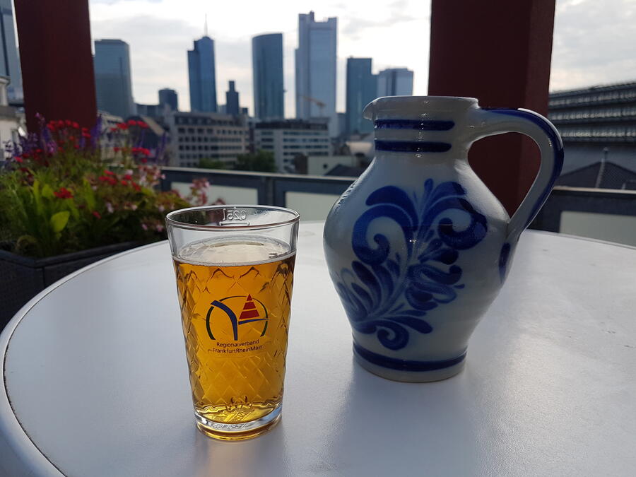 Ein typisch hessisches Apfelweinglas "Geripptes", daneben ein Bembel, im Hintergrund sieht man die Skyline von Frankfurt.