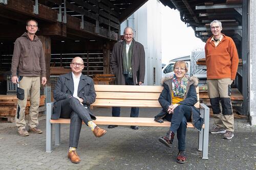 Fünf Personen stehen und sitzen vor einer Holzlagerhalle.